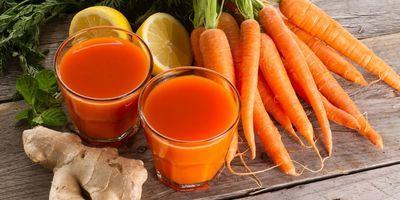 2 glasses containing a fresh carrot smoothie. Fresh carrots, lemons and ginger root surround the glasses.